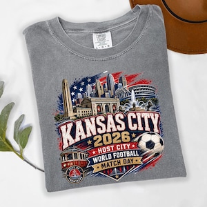 May include: A grey crew neck t-shirt featuring a graphic print of the Kansas City skyline, a red streetcar, and a soccer ball. The text reads KANSAS CITY 2026 HOST CITY WORLD FOOTBALL MATCH DAY. The shirt has a Comfort Colors brand label at the neck.
