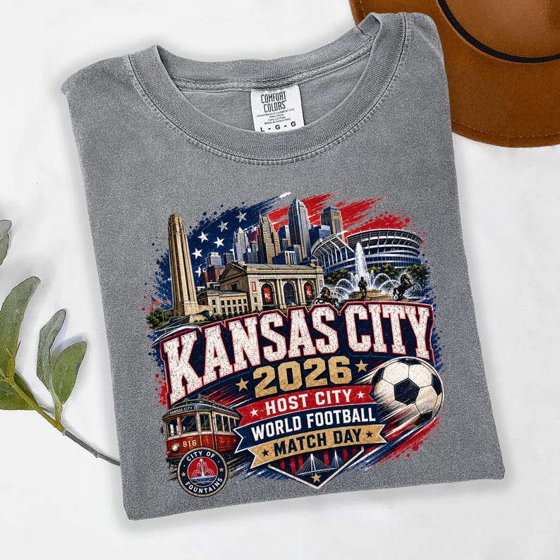 May include: A grey crew neck t-shirt featuring a graphic print of the Kansas City skyline, a red streetcar, and a soccer ball. The text reads KANSAS CITY 2026 HOST CITY WORLD FOOTBALL MATCH DAY. The shirt has a Comfort Colors brand label at the neck.