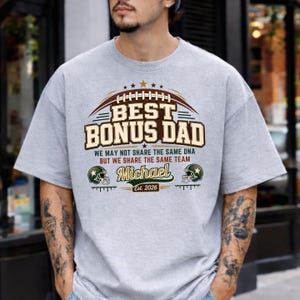 May include: A light gray t-shirt with the text "BEST BONUS DAD" in a football-themed design. The shirt also includes the name "Michael" and the year "Est. 2026". The design features football helmets and stars.