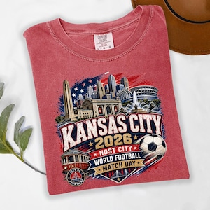 May include: A red short-sleeve t-shirt featuring a graphic of the Kansas City skyline, a streetcar, and a soccer ball. The text reads KANSAS CITY 2026 HOST CITY WORLD FOOTBALL MATCH DAY and CITY OF FOUNTAINS. The shirt has a Comfort Colors brand label.