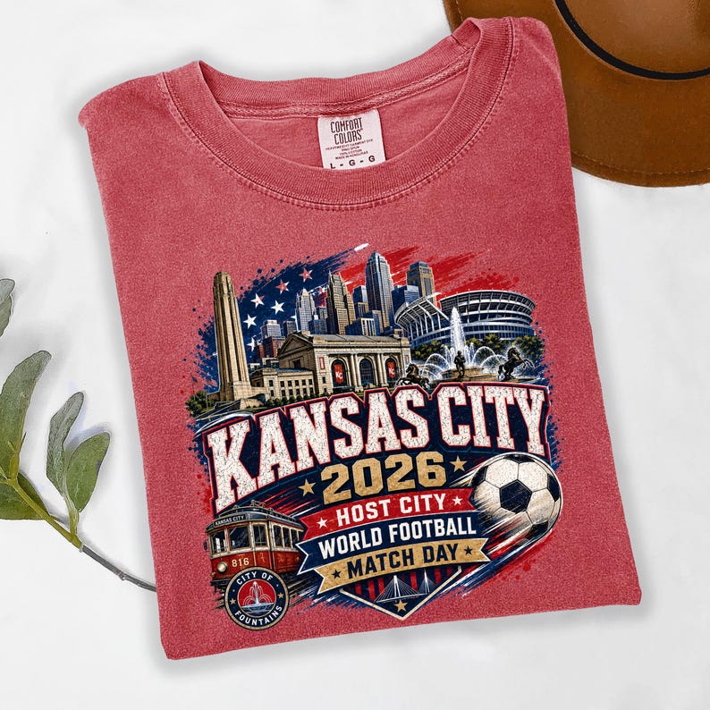 May include: A red short-sleeve t-shirt featuring a graphic of the Kansas City skyline, a streetcar, and a soccer ball. The text reads KANSAS CITY 2026 HOST CITY WORLD FOOTBALL MATCH DAY and CITY OF FOUNTAINS. The shirt has a Comfort Colors brand label.