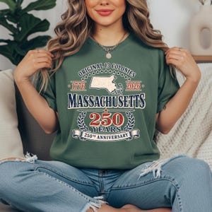 May include: Green t-shirt with the text "Original 13 Colonies" and "Massachusetts 250 Years 250th Anniversary". The design includes a map of Massachusetts, the years 1776 and 2026, and a banner.