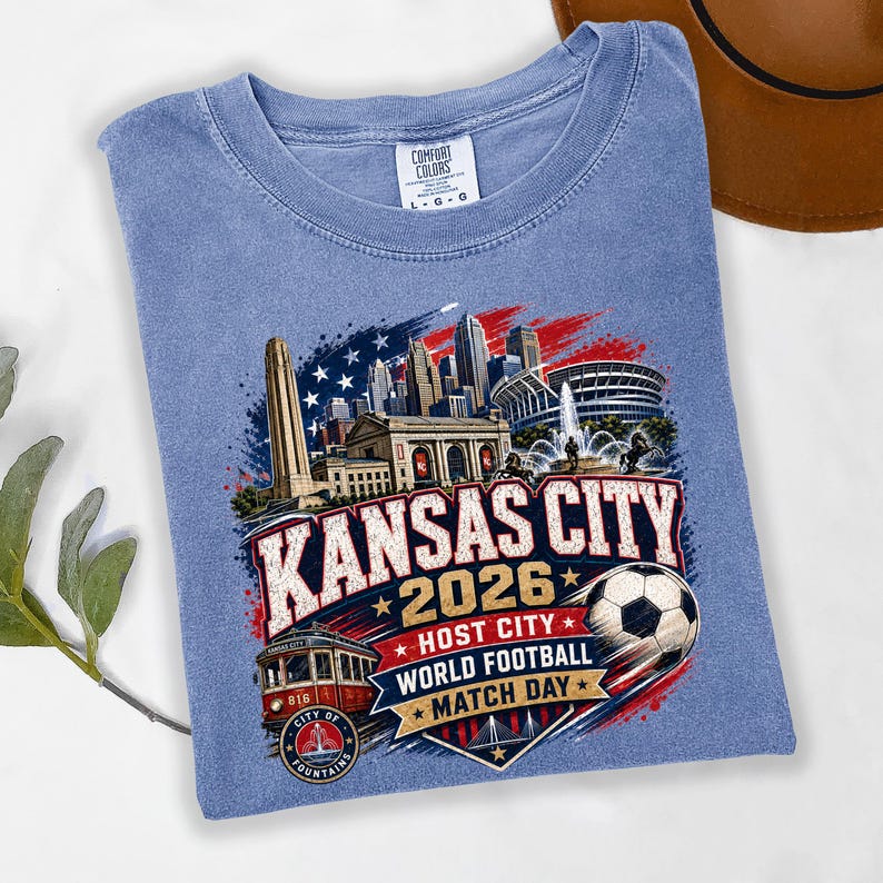 May include: A blue crew neck t-shirt featuring a graphic of the Kansas City skyline, a red streetcar, and a soccer ball. The text reads Kansas City 2026 Host City World Football Match Day. The shirt has a Comfort Colors label indicating size large.