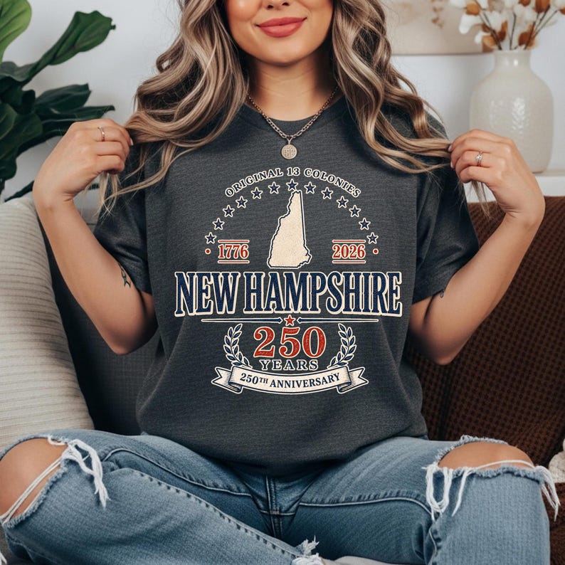 May include: Dark gray t-shirt with a graphic celebrating the 250th anniversary of New Hampshire. The design includes the state outline, the text "NEW HAMPSHIRE", and the years 1776 and 2026. The shirt also features the text "ORIGINAL 13 COLONIES".