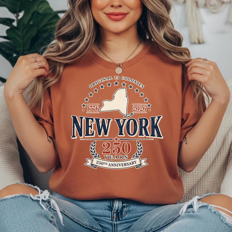 May include: A rust-colored t-shirt featuring a design commemorating the 250th anniversary of New York. The design includes the text "Original 13 Colonies," the New York state outline, and the years 1776 and 2026.