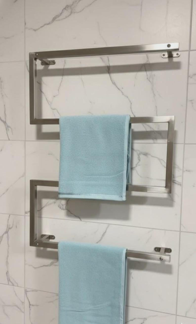 May include: A stainless steel towel rack mounted on a tiled wall, holding two light blue towels. The rack has a modern, geometric design with three horizontal bars for hanging towels. The wall tiles have a marble-like pattern.