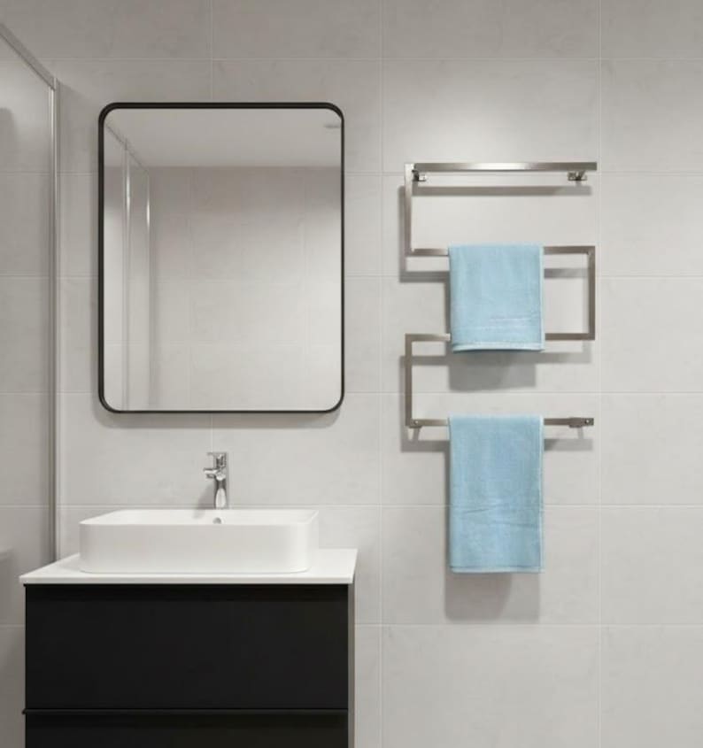 May include: A modern bathroom scene featuring a rectangular mirror with a black frame, a white rectangular sink atop a black cabinet, and a stainless steel towel rack with two light blue towels. The walls are tiled in a light gray color.