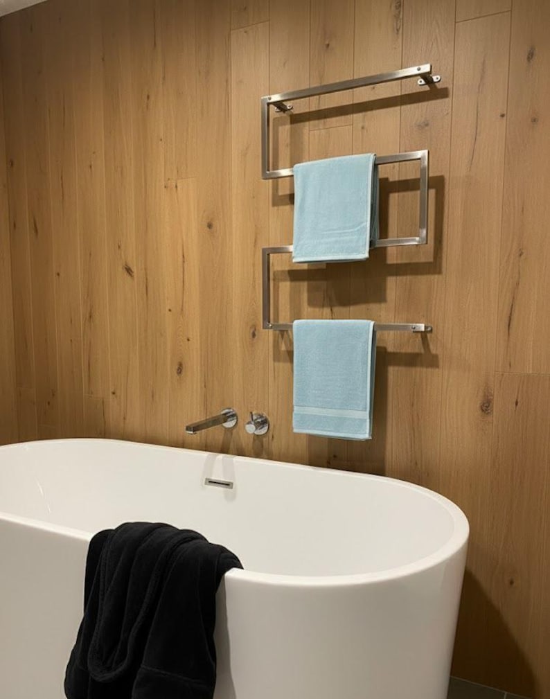 May include: A modern bathroom scene featuring a white oval bathtub and a stainless steel towel rack with two light blue towels. The wall is paneled with light brown wood. A black robe is draped over the tub's edge.