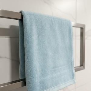 May include: A light blue bath towel hanging on a stainless steel towel rack. The towel has a textured surface and a decorative border. The background is a white tiled wall.