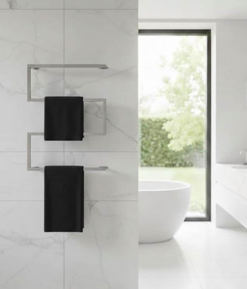 May include: A modern, stainless steel towel warmer with two black towels hanging on it. The towel warmer is mounted on a white tiled wall. The bathroom has a large window and a white bathtub.
