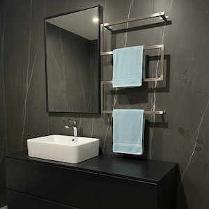 May include: A modern bathroom scene featuring a black vanity with a white rectangular sink and a chrome faucet. A rectangular mirror with a black frame hangs above the sink. A stainless steel towel warmer holds two light blue towels. The walls are dark gray with white veining.
