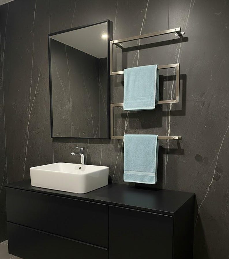 May include: A modern bathroom scene featuring a black vanity with a white rectangular sink and a chrome faucet. A rectangular mirror with a black frame hangs above the sink. A stainless steel towel warmer holds two light blue towels. The walls are dark gray with white veining.