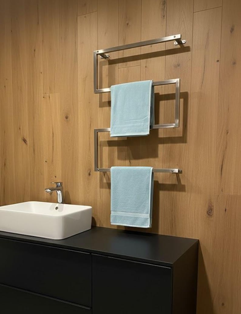May include: A modern bathroom scene featuring a stainless steel towel rack with three levels, each holding a light blue towel. The rack is mounted on a wooden wall above a black cabinet with a white rectangular sink and chrome faucet.