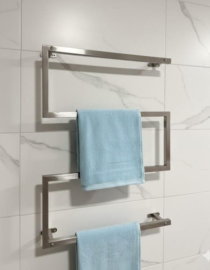 May include: A modern, stainless steel towel warmer mounted on a white tiled wall. The geometric design features three horizontal bars for hanging towels. Two light blue towels are neatly displayed on the warmer, adding a pop of color to the bathroom.