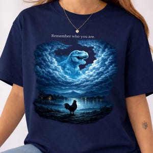 Puede incluir: Camiseta azul marino con un diseño surrealista. Presenta un Tyrannosaurus Rex azul en las nubes, un gallo y un paisaje lacustre. El texto "Remember who you are" está encima de la imagen.