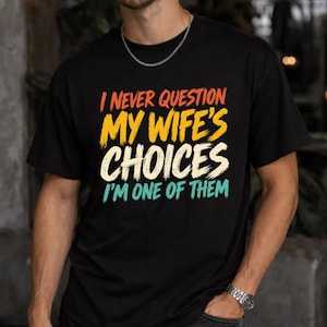 May include: A black short-sleeved t-shirt featuring the text I NEVER QUESTION MY WIFE'S CHOICES I'M ONE OF THEM in a distressed, multi-coloured font. The words are stacked in orange, yellow, cream, and teal, creating a bold graphic design.