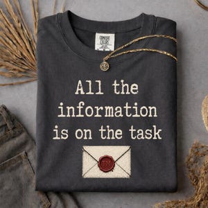 May include: Dark grey Comfort Colors t-shirt with the text "All the information is on the task" in white. An illustration of an envelope with a red wax seal is below the text. A gold necklace with a pendant is on the shirt.