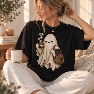 May include: A black t-shirt featuring a graphic of a white ghost holding a brown mug with the text "MEH" and a heart-shaped steam trail. The ghost carries a brown bag containing a small black pug. The design is centered on the front of the shirt.