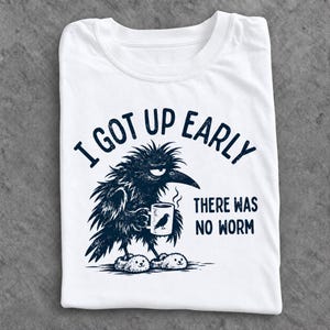 Könnte beinhalten: Ein weißes T-Shirt mit einer dunkelblauen Grafik eines zerzausten Raben, der flauschige Hausschuhe trägt und eine dampfende Tasse hält. Der Text lautet I GOT UP EARLY THERE WAS NO WORM in einer abgenutzten Schriftart mit Rabensilhouette.