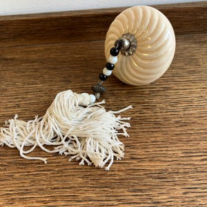 May include: A cream-coloured ceramic knob with a swirling, ribbed pattern. The knob features a decorative attachment consisting of alternating black and white beads, a small metallic spacer, and a long, off-white fabric tassel with loose threads.