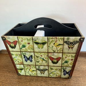 May include: A square decorative storage bin with a black handle. The bin features a collage of butterflies and birds on a light green background. The edges are trimmed with brown and metal corner accents.