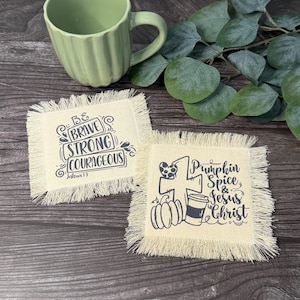 May include: Two square fabric coasters with frayed edges. One coaster features the black text Be Brave Strong Courageous Joshua 1:9. The other coaster displays a cross, a pumpkin, and a coffee cup with the text Pumpkin Spice & Jesus Christ.