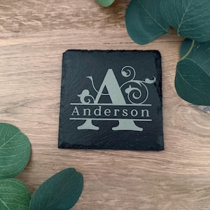 May include: A square black slate coaster featuring an engraved design with a large capital letter A, decorative floral swirls, and the name Anderson printed in a horizontal line across the center of the letter in a serif font.