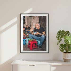 May include: A framed photographic print featuring a man with gray hair wearing a black t-shirt and blue jeans, sitting on a small red plastic stool while eating with chopsticks from a bowl. Two other individuals are visible in the background.
