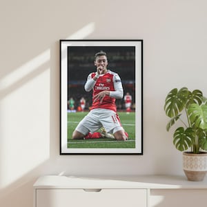 May include: Framed print featuring a soccer player in a red and white jersey, kneeling on a green field. The player is celebrating with a hand gesture. The frame is black, and the print is displayed on a white surface.