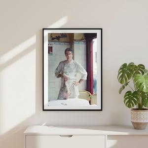 Può includere: Una stampa fotografica incorniciata che ritrae uno chef con una giacca bianca a doppio petto e un grembiule, mentre tiene in mano un pezzo di carne cruda. Lo chef si trova in una cucina con pareti piastrellate chiare, una porta scura e un tavolo.