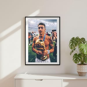 May include: Framed photograph of a soccer player holding the World Cup trophy. The player is shirtless, wearing white shorts, and a blue medal. The trophy is gold and the background shows a stadium with spectators.