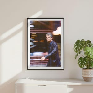May include: A framed photographic print featuring a man with gray hair wearing a dark blazer and blue shirt. The image has a motion blur effect with warm light streaks in the background, presented in a thin black frame on a plain wall.