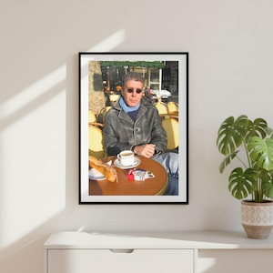 May include: A framed photograph of a person wearing a dark leather jacket, a light blue scarf, and round sunglasses, sitting at a round outdoor cafe table with a baguette, a white coffee cup, and a pack of cigarettes on the wooden surface.