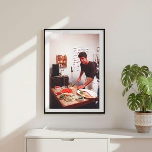 May include: A framed photograph of a person in a dark long-sleeved shirt and white apron preparing food on a wooden kitchen island. The scene includes a black microwave, various small bowls of ingredients, a fish on a platter, and a wall chart.