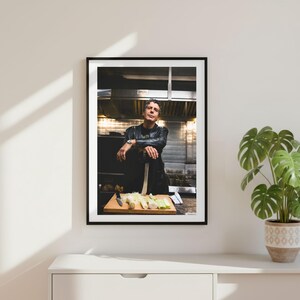 Può includere: Una stampa fotografica incorniciata che ritrae una persona con una giacca di pelle nera in una cucina professionale. La persona appoggia le braccia su un tagliere di legno con porri affettati e un coltello, su uno sfondo in acciaio inox.