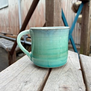 Puede incluir: Taza de cerámica hecha a mano con un esmalte degradado en tonos turquesa y verde. La taza tiene un cuerpo redondeado, un asa curvada y una superficie ligeramente texturizada. Está sobre una superficie de madera.