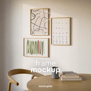 Puede incluir: Tres impresiones artísticas enmarcadas con diseños abstractos cuelgan de una pared blanca. Las impresiones presentan un fondo beige con líneas negras, líneas verticales verdes y rojas y formas azules. Una mesa y una silla de madera están en primer plano, con el texto "frame mockup" y "mockupize".