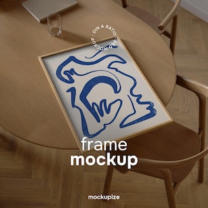 May include: A framed art print featuring a blue abstract face design on a cream background. The frame is light brown, and the artwork is displayed on a wooden table with a glass and a laptop. The words "frame mockup" are printed on the frame.