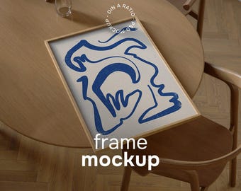 May include: A framed art print featuring a blue abstract face design on a cream background. The frame is light brown, and the artwork is displayed on a wooden table with a glass and a laptop. The words "frame mockup" are printed on the frame.
