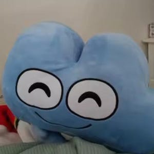 May include: A large, light blue plush toy shaped like a stylized number four. It has two large, white-rimmed eyes with black curved pupils and a smiling mouth. The plush is resting on a green blanket.