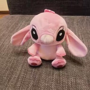 May include: A pink plush toy of a cartoon character with large ears, black eyes, and a small nose. The toy has a small tuft of hair on its head and is sitting on a textured surface.