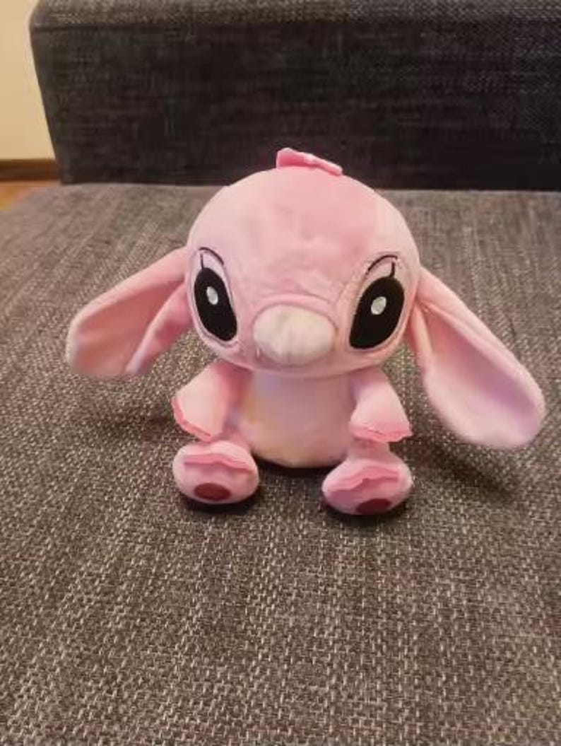 May include: A pink plush toy of a cartoon character with large ears, black eyes, and a small nose. The toy has a small tuft of hair on its head and is sitting on a textured surface.