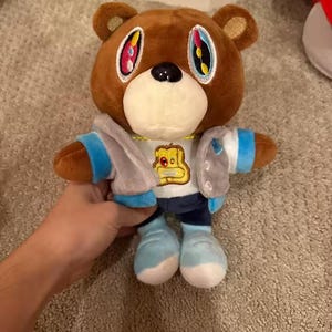 May include: Brown plush bear wearing a light blue and grey jacket, white shirt with a gold graphic, and blue trousers and shoes. The bear has large, colourful eyes and a white muzzle. A collectible plush toy.