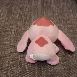 May include: A pink plush toy with large ears and a red patch on its back. The toy is lying on a textured brown surface. The plushie has a white tag on its bottom.