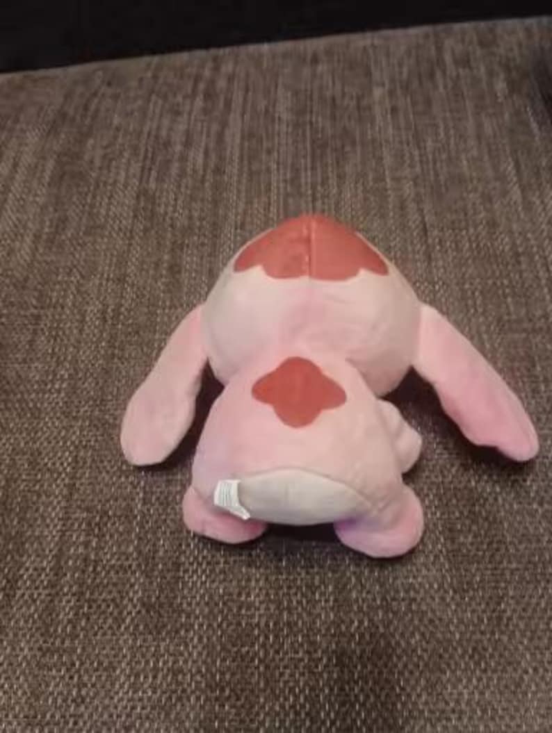 May include: A pink plush toy with large ears and a red patch on its back. The toy is lying on a textured brown surface. The plushie has a white tag on its bottom.
