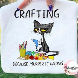 May include: White t-shirt featuring the word "CRAFTING" and a cartoon cat with a glue gun. The text "BECAUSE MURDER IS WRONG" is below the cat. Crafting supplies such as paint and yarn are also visible.