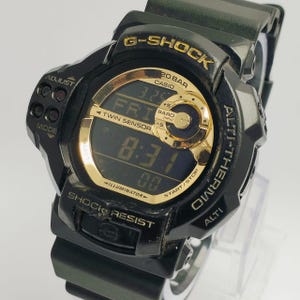 Puede incluir: Reloj digital G-Shock negro con detalles dorados. El reloj muestra la hora, la fecha y otras funciones en una pantalla digital. Tiene botones para ajustar el modo, iniciar/detener y otras funciones. El reloj es resistente a los golpes e incluye un altímetro y un termómetro.