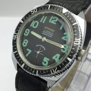 May include: A vintage Sphinx wristwatch with a black face and luminous green numerals. The watch features a rotating bezel, a black leather band, and the text "ANTIMAGNETIC". The watch is marked "17 JEWELS WATERPROOF SHOCKPROOF".
