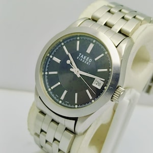 May include: A silver-toned TAKE KIKUCHI wristwatch with a dark gray face and a date display at the 3 o'clock position. The watch has a stainless steel band and a textured bezel. The watch is displayed on a white surface.