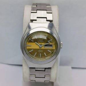 May include: A vintage Citizen automatic wristwatch with a silver-tone metal band and case. The watch face is gold with a horizontal stripe and displays the day and date. The watch is set on a white display stand.